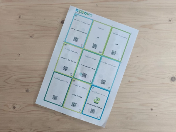 Photo d'une feuille de cartes-jeu Koolookoo posée sur une table en bois, avec des cartes QR code colorées portant des titres d'exercices (Voyelles et consonnes, Frappe de phrase, Coloriage guidé, Principe des syllabes, etc.).